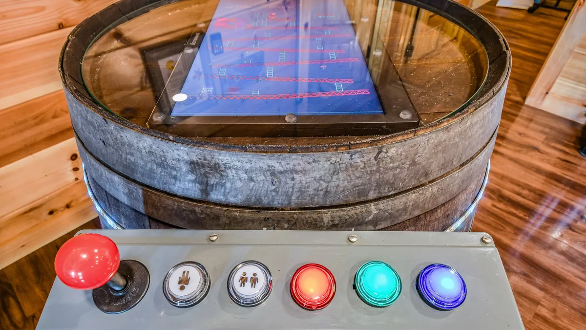 An interactive arcade table sits at the center of a rustic room, complete with a barrel-shaped base, touchscreen, and colorful control buttons, inviting guests to relive classic games and engage in nostalgic fun.