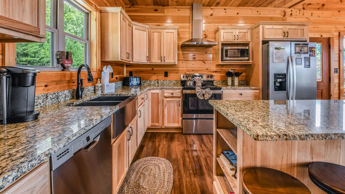A modern chef’s kitchen equipped with granite countertops, wooden cabinetry, stainless steel appliances, and a central island. Large windows flood the space with light, offering pleasant views while preparing meals or gathering around the island.