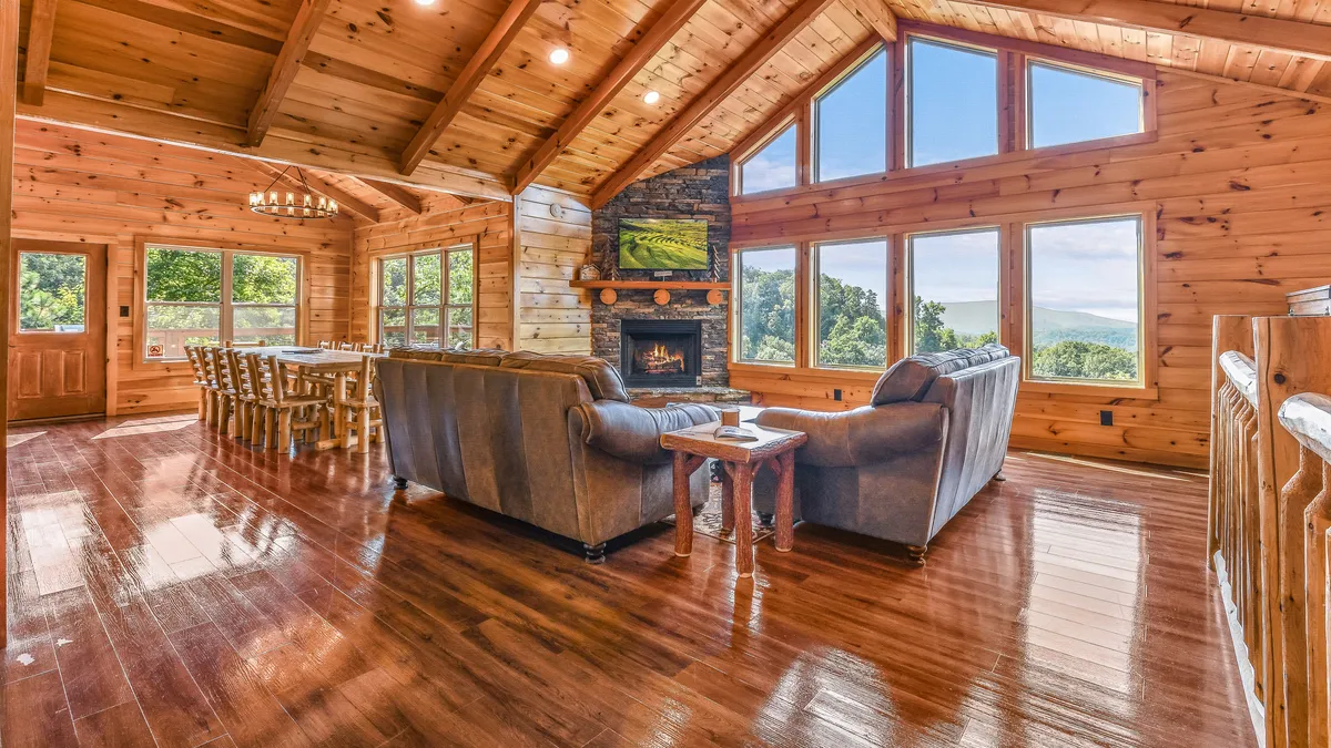 A spacious open-concept living and dining area, featuring large windows that reveal stunning mountain views. The cozy stone fireplace is the centerpiece, surrounded by comfortable sofas and a substantial wooden dining table. Warm wood beams and natural light enhance the welcoming ambiance, inviting group gatherings and relaxation.