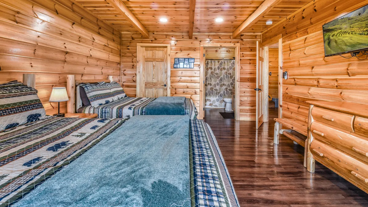 A guest suite with multiple beds, decorative bedding, a wall-mounted television, and an en suite bathroom door. The warm wooden paneling and practical layout invite relaxation and group stays.