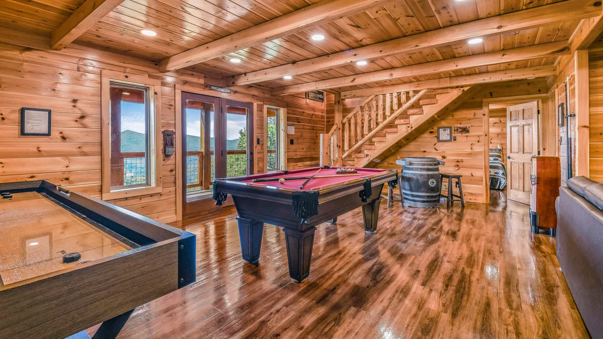 This image showcases a spacious game room with a pool table and foosball table at the center, large windows offering scenic mountain views, and a wooden staircase leading to the space. The room's warm lighting and wood finishes create a cozy, playful atmosphere ideal for gatherings and friendly competitions.