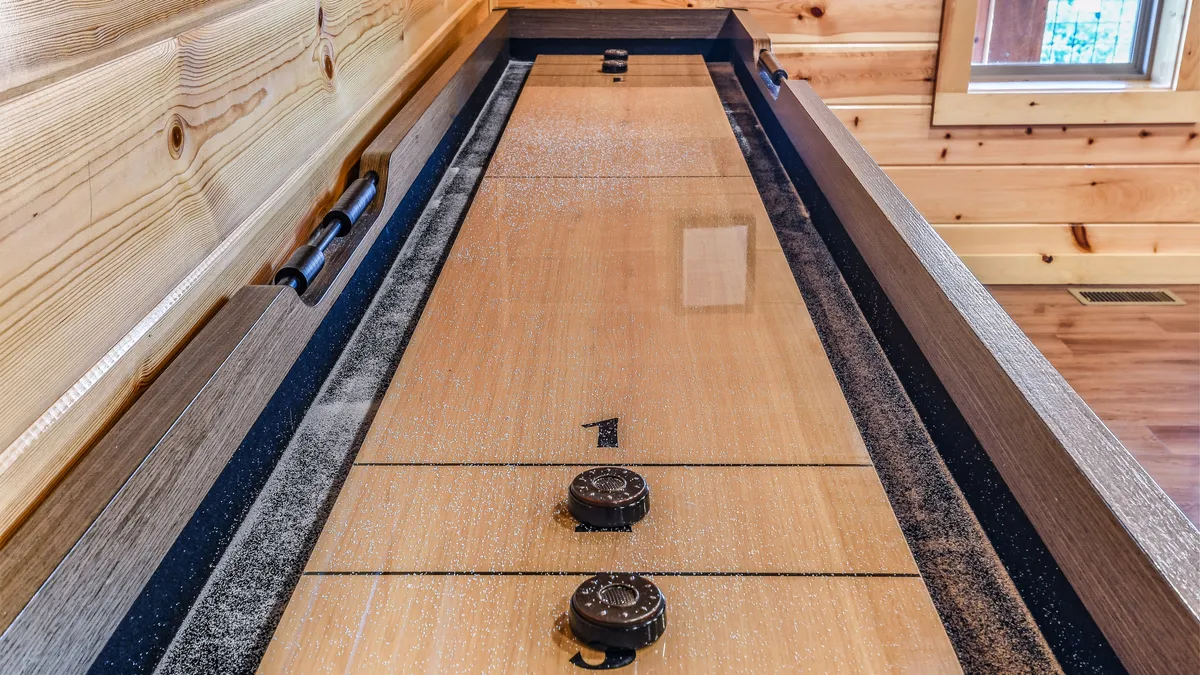 A shuffleboard table runs along one side of a warmly lit game area, lined with natural wood paneling and bathed in natural light from a nearby window. The scene suggests a space designed for playful competition and casual gatherings.