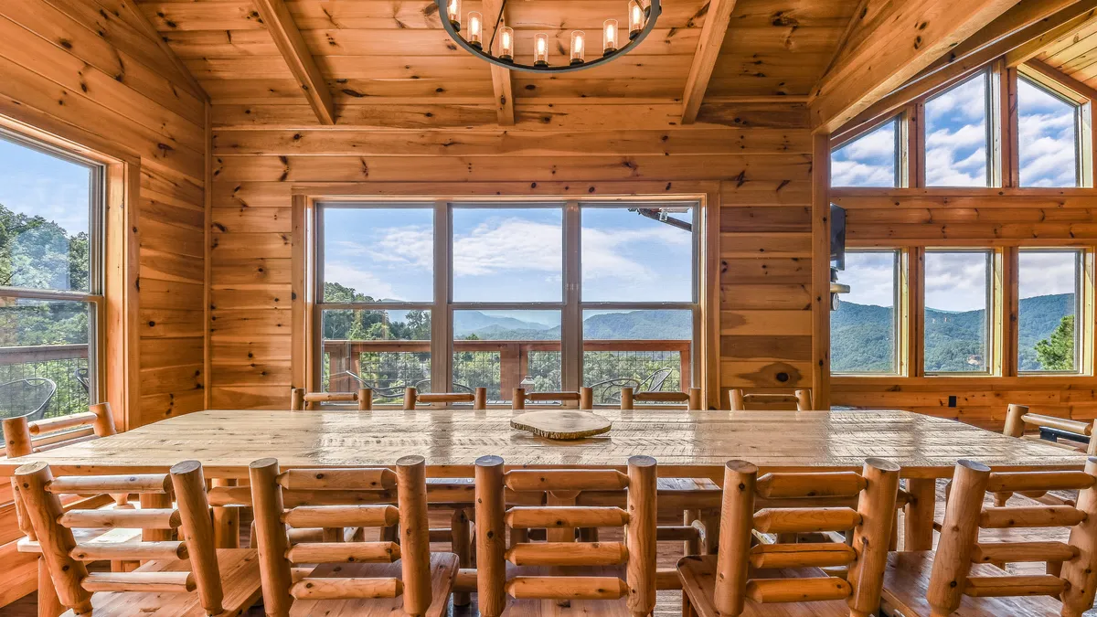 A long rustic wooden dining table set beneath a striking chandelier, perfectly positioned next to floor-to-ceiling windows that showcase the outdoors. The arrangement is ideal for large groups, and the ample natural light creates an inviting atmosphere for memorable meals and celebrations.