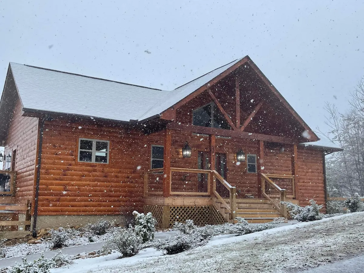 A picturesque log cabin facade with a covered front porch and snow-dusted roof, illuminated by warm lanterns. The scene radiates coziness and is set against a winter backdrop, highlighting the cabin's charm during colder months and invoking feelings of warmth and welcome.
