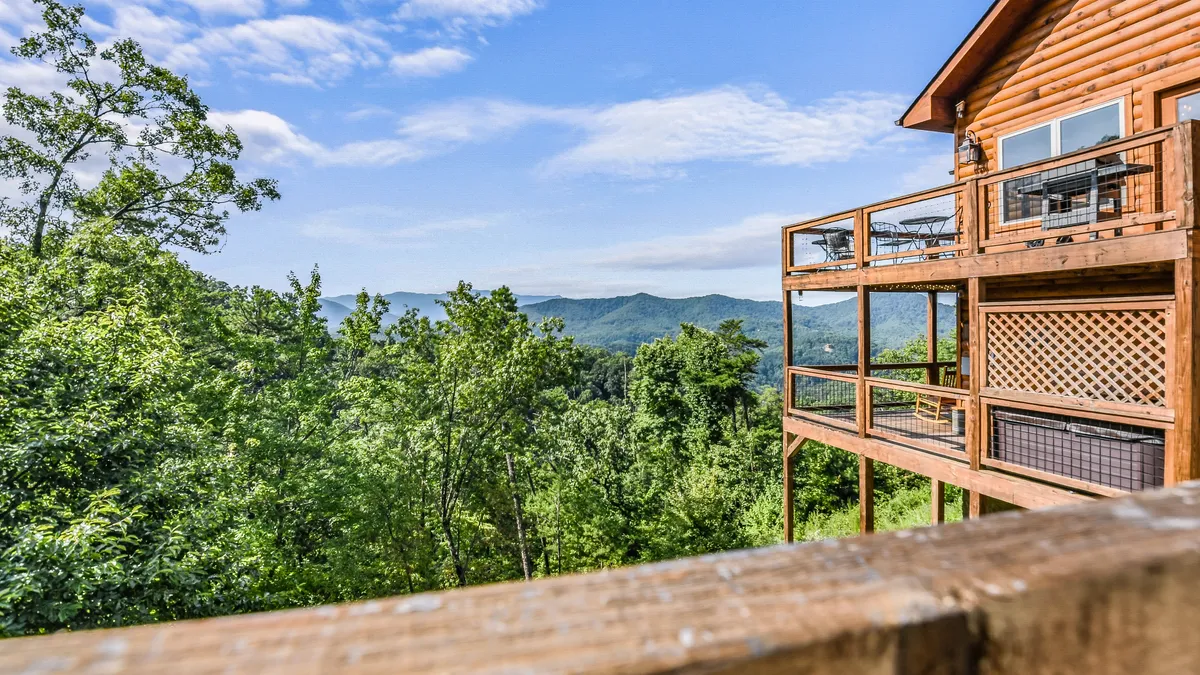 A spacious wooden deck with comfortable seating overlooks lush greenery and rolling mountains. The deck is surrounded by a sturdy railing and provides an expansive view, inviting guests to relax with a morning coffee or stargaze at night. Sunlight casts a warm glow over the scene, highlighting the peaceful natural setting.