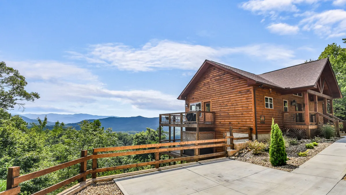 The photo shows a welcoming log cabin with a wooden deck, surrounded by greenery and mountain views, representing the cabin's proximity to outdoor adventures and its inviting atmosphere for returning after a day of exploration.