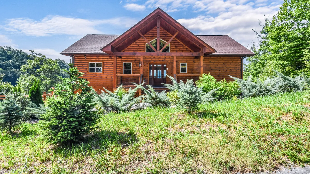 This image features a classic log cabin exterior with a large front porch surrounded by green landscaping. The inviting architecture hints at group-friendly gatherings and outdoor relaxation, perfect for welcoming guests.